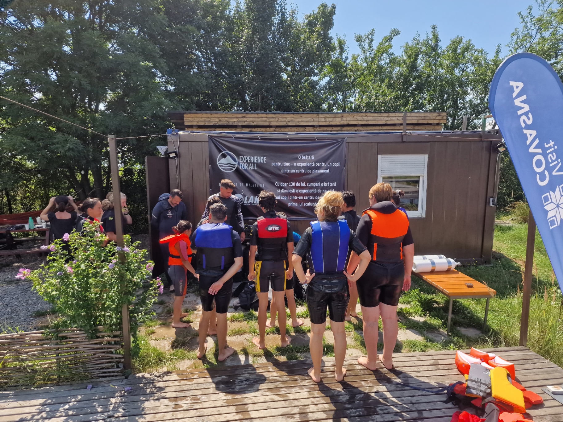 Participanți în costume de neopren și veste de salvare, adunați lângă bannerul Experience for All la baza de pe lac