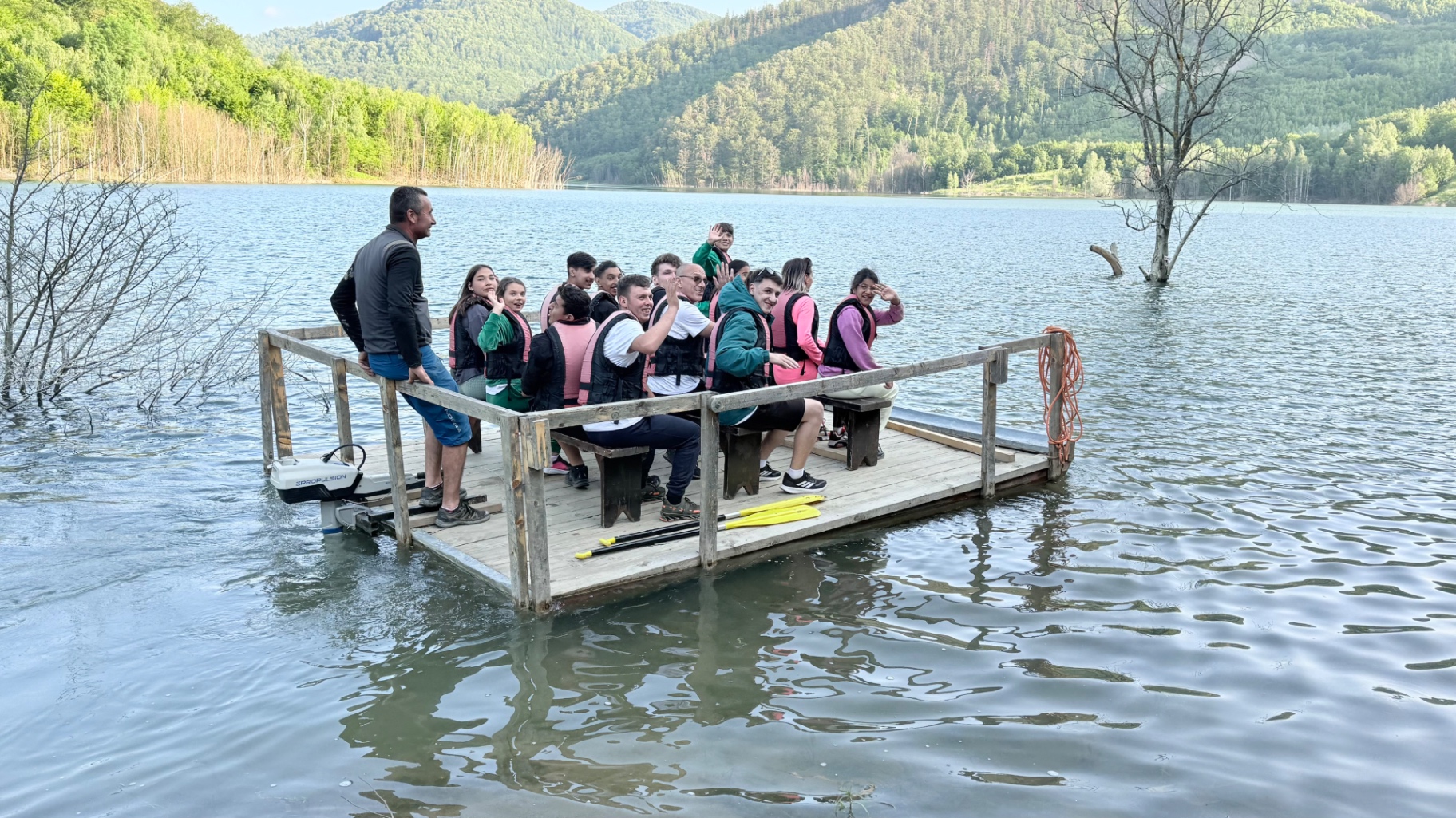Grup de adolescenți cu veste de salvare pe o plută de lemn, pe un lac înconjurat de dealuri împădurite