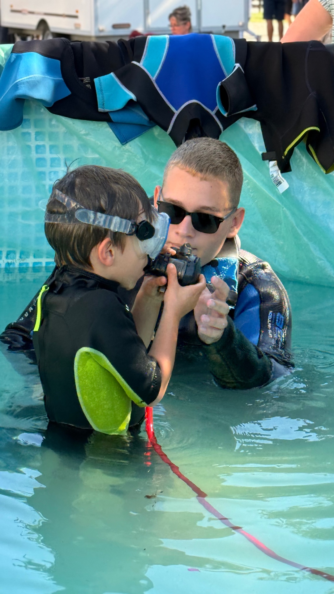 Copil în costum de neopren care respiră prima dată pe regulator, sprijinit de un instructor, într-o piscină